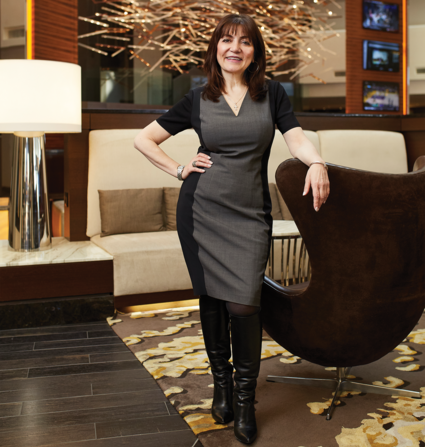 Vera Manoukian rests her arm on a black leather chair while posing for a portrait in an upscale hotel setting.