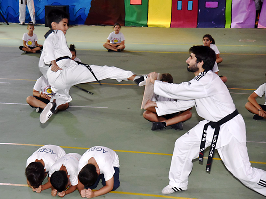 A martial arts test where one child leaps over 3 others are laying on the ground to kick a board held by an instructor. 