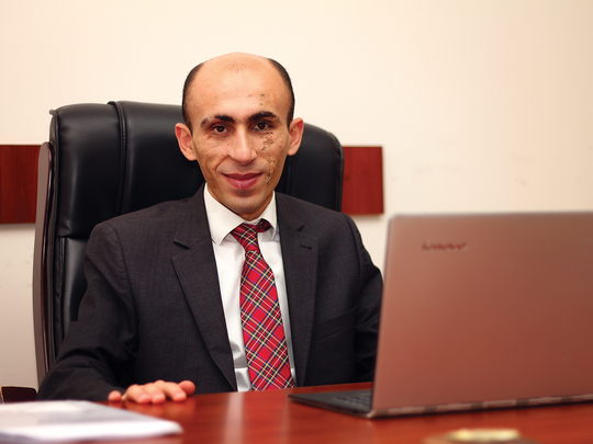 A man in a black suite, white shirt and a red tie sitting on his desk in front of his computer