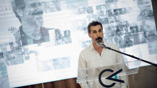 Serj Tankian standing at a podium with his hands in his pockets in front of a presentation slide